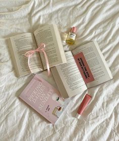 an open book, lip glosses, and perfume on a bed with white sheets