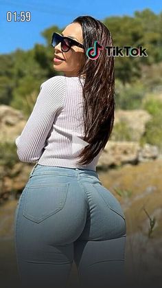 a woman with long hair and sunglasses standing in front of a body of water wearing high waist jeans