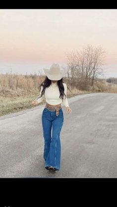 a woman walking down the middle of a road wearing jeans and a cowboy hat on her head