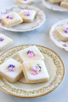 some white and purple decorated cookies are on plates with pink flowers in the middle, along with other small ones