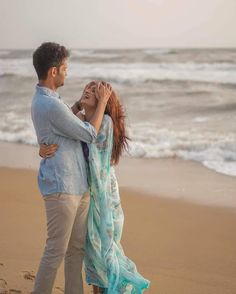 a man and woman standing on top of a sandy beach next to the ocean holding each other