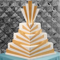 a three tiered cake with gold and white stripes on the top, sitting on a blue table cloth