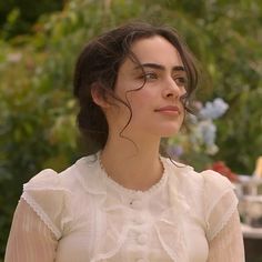 a woman in a white dress is looking off to the side with trees and bushes behind her