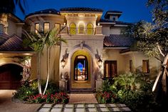 the entrance to a large home at night with palm trees and flowers in front of it