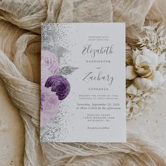 a wedding card with purple flowers on it next to some white flowers and burlock