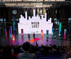 two men standing on stage in front of an audience with the moon shut screen behind them