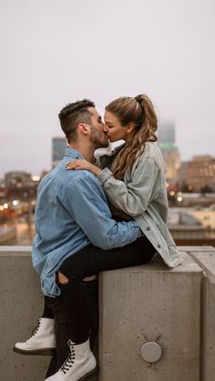 a man and woman kissing while sitting on top of a cement wall in front of a cityscape