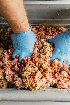 two hands in blue gloves scoop food out of a metal pan into a bin filled with meat