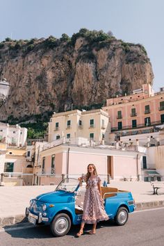 a woman standing next to a blue car in front of some buildings and a mountain