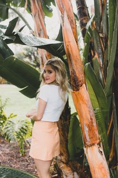 a woman standing next to a tree with lots of green leaves on it and looking at the camera