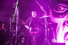 a man playing drums on stage with purple lighting behind him and an evil demon painted on the wall behind him