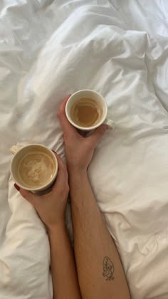 two cups of coffee sitting on top of someone's legs in bed with white sheets