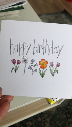 a person holding up a card with flowers on it that says happy birthday written in black ink