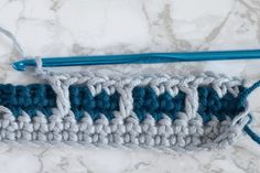 the crochet stitch is being worked on by a blue handled knitting needle, along with two rows of white and gray yarn