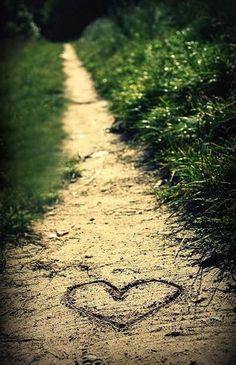 a small heart drawn in the sand on a dirt path near grass and bushes,