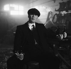 a man in a suit and hat holding two beer bottles while sitting on a chair