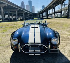 an old blue and white sports car parked in front of a highway overpass with skyscrapers in the background