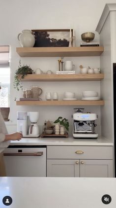 a woman standing in front of a kitchen counter filled with dishes and coffee cups on top of wooden shelves