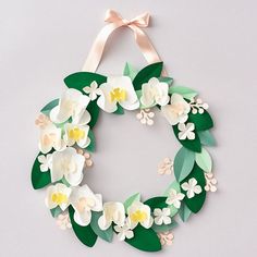 a paper wreath with white flowers and green leaves hanging from a ribbon on a gray background