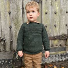 a little boy standing in front of a wooden fence wearing a green sweater and khaki pants