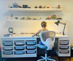 a young boy sitting at a desk with toy cars on the shelves above him and toys in bins behind him