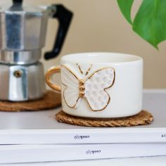 a white coffee cup with a butterfly on it sitting next to a silver teapot