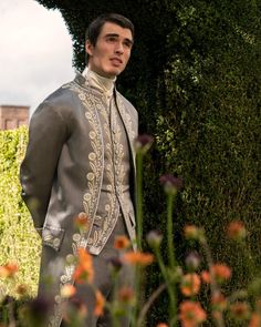 a man dressed in an elaborately designed suit standing next to a bush and flowers