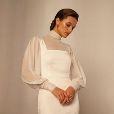 a woman wearing a white dress with sheer sleeves and high heels standing in front of a wall