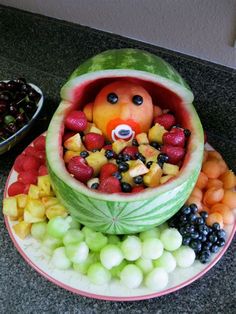 a watermelon bowl filled with fruit and a teddy bear in the center surrounded by other fruits