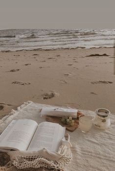 an open book sitting on top of a sandy beach next to the ocean with food