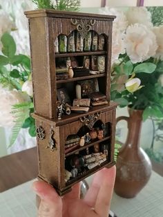 a person holding up a miniature wooden bookcase with many items on it and flowers in the background