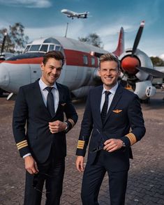 two men in suits standing next to each other near an airplane and another plane behind them