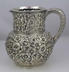 an ornate silver pitcher with floral designs on the outside and inside, sitting on a white surface