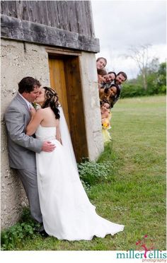 a bride and groom kissing in front of an outhouse with their wedding party behind them