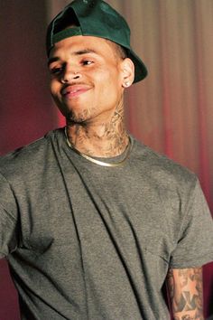 a man with tattoos wearing a hat and looking at the camera while standing in front of a curtain