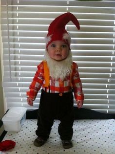 a baby wearing a red and white gnome hat standing on top of a bed next to a window