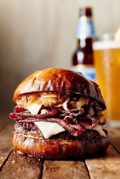 a sandwich with meat and cheese on a wooden table next to a glass of beer