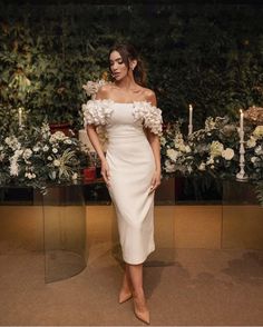 a woman in a white dress standing next to some candles and flowers on the wall