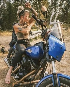 a woman sitting on top of a blue motorcycle in the dirt with trees behind her