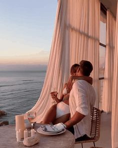 a man and woman sitting at a table next to the ocean with wine glasses in their hands