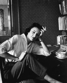 a black and white photo of a woman sitting in front of a book shelf holding her hand to her head