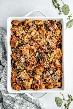 a casserole dish filled with stuffing and vegetables on a white plate next to some herbs