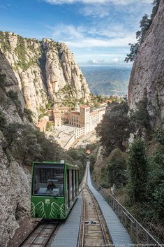 a green train traveling down tracks next to a mountain side town and forest covered mountains