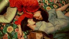 two people laying on a bed with flowers in the background and one person wearing a red shirt