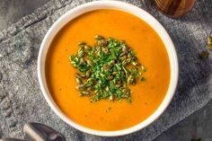 a white bowl filled with carrot soup and topped with pistachio seeds, garnished with chopped green leaves