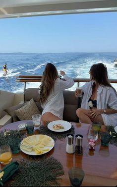 two women sitting at a table with food and drinks in front of an ocean view