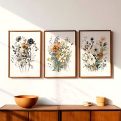 three framed paintings on the wall above a wooden cabinet with a bowl and cup sitting on it