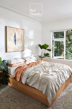 an unmade bed in a white room with plants on the floor and a painting hanging above it