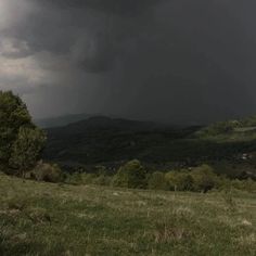 a dark cloud hovers in the sky over a grassy field with trees and hills