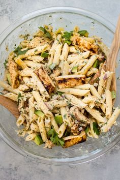 a glass bowl filled with pasta and chicken on top of a gray counter next to a wooden spoon